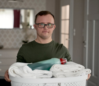 A man wearing glasses and an olive drab shirt holds a white laundry basket filled with white towels and a bottle of laundry detergent indoors. The setting is a well-lit room, likely a home, with a white dresser, framed mirror and door in the background. The composition is a waist-up shot, capturing the man's upper body and the laundry basket. The mood is calm and domestic. This image could be used for advertising home products, illustrating concepts of cleanliness and responsibility, or representing inclusive lifestyle content.
