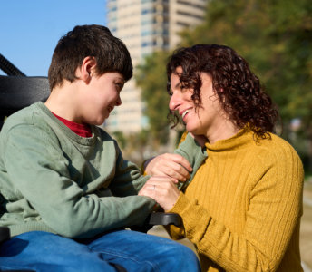 A smiling child in a wheelchair is holding hands with an adult in an urban park setting. The child has dark hair and is wearing a green sweater and blue jeans. The adult has wavy dark hair and is wearing a yellow sweater. The subjects are smiling at each other. The mood of the photo is uplifting. This photograph could be used for projects centered on care, family, connection, and outdoor moments. The photograph could also be used for projects related to disabilities.