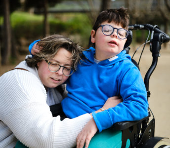 In a tranquil outdoor setting, a woman with short, wavy brown hair and wearing glasses affectionately embraces a child in a wheelchair. The child, who is wearing glasses and a blue hoodie, sits relaxed in their chair. The woman is wearing a white sweater, and has a backpack. The scene evokes a mood of connection, peace, and warmth, suggesting themes of support and affection. The photo could be used for projects related to caregiving, special needs, family moments, or accessible outdoor activities. It could also illustrate themes of unconditional love.
