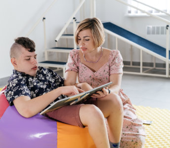 A teenager with dark, textured hair and a floral shirt sits on a colorful beanbag chair with an adult in a floral dress. The adult points at a book that the teenager is looking at, in a well-lit room with exercise equipment. This serene image captures a moment of shared reading, making it ideal for projects related to education, therapy, and home activities. The soft lighting enhances the calm mood, suitable for conveying concepts of guidance and development.