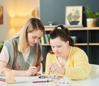 Two young women are seated at a table, creating art together in a brightly lit interior. The woman on the left, with long blonde hair, wears a pale pink t-shirt with a green, pink, and grey vest. She is holding a paintbrush, focused on the artwork in front of her. The woman on the right, with dark hair pulled back, wears a yellow sweater and is similarly intent on the art project. The setting appears to be a home with shelves, plants, and art on the walls. This image could be used in projects about friendship, creative expression, or art instruction.