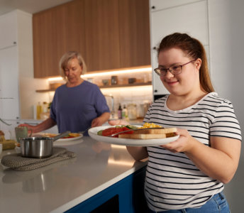 A smiling young woman with auburn hair and glasses holds out a plate with toast, scrambled eggs, sliced tomatoes, and cucumbers, looking directly at the camera. She wears a white t-shirt with black horizontal stripes. In the background, a woman with short blonde hair stands near the stove. The scene is set in a bright, modern kitchen with light wood cabinets. The photo's bright lighting and natural composition create a welcoming mood. This image would be well-suited for illustrating healthy eating, family life, or cheerful mornings.