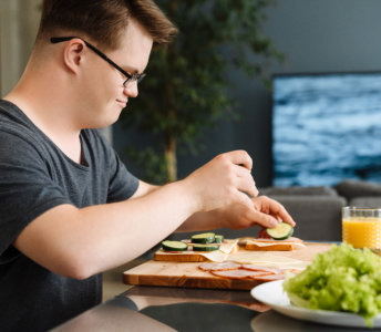 A man with short brown hair and wearing glasses is making sandwiches at a counter. He's adding sliced cucumbers on top of cheese and ham. In the foreground sits a plate of lettuce and a glass of orange juice. The photo is brightly lit with natural light, and the setting looks like a kitchen or dining area inside a home. This photo would be great for content about healthy eating, cooking at home, family activities, and wellness.