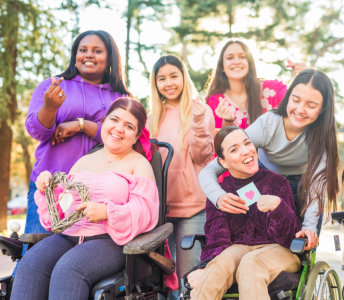 A group of eight women smile outdoors in a park. Three of the women are in wheelchairs. Most of the women are wearing pink or purple clothing. One woman in the center of the shot is holding a card with a pink heart on it. Some of the women are holding their hands in the shape of a heart. This scene is perfect for conveying themes of friendship, support, and care. Use it for projects about community, connection, and helping others.