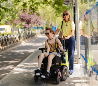 A cheerful young woman with reddish hair in a wheelchair and a woman with blonde hair stroll along an urban sidewalk near a bus stop on a bright sunny day. The young woman wears a patterned top, denim shorts, and sunglasses, and is smiling. The older woman is wearing a yellow top, jeans, and sunglasses, and pushes the wheelchair along a sidewalk in a vibrant urban setting with lush green trees lining the street. The photo's bright lighting and natural setting create a light-hearted mood, making it suitable for projects related to lifestyle, accessibility, family, support, and outdoor activities.