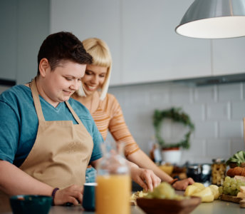 Two adults are visible in this bright, airy photo, standing in a kitchen. One adult wears a beige apron and is cutting fruit on a countertop. A second adult with blonde hair is behind them, smiling. Bowls of fruit, prepared food, and a jar of orange juice are visible. The scene has a light, carefree mood. This image could be used in projects related to food preparation, family, healthy eating, togetherness, and home life.