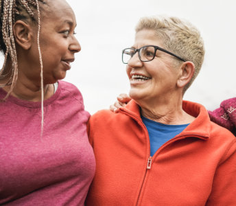This bright, cheerful photo shows two women standing close together. One woman has blonde braided hair and wears a pink t-shirt. The other woman wears glasses and an orange fleece jacket. Both women are smiling and in an urban setting. The photo's lighthearted mood makes it ideal for campaigns related to friendship, family, or health and wellness. The image is in a waist up perspective.
