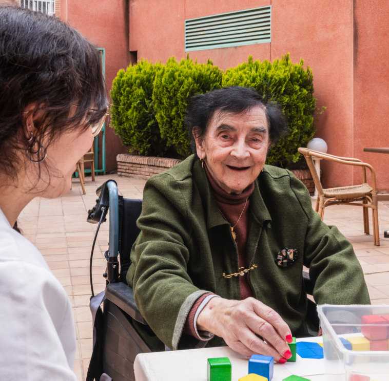 A kind caretaker assists a senior woman as she plays with colorful blocks on an outdoor patio. The woman, seated in a wheelchair, smiles gently and wears an olive green jacket. The caretaker, facing away from the camera, has dark curly hair. They are outside on a brick paved patio with a red building and green door in the background. This image could be used for projects centered on care facilities, wellness, senior care, rehabilitation, or assisted living. It also conveys themes of connection, compassion, and support.