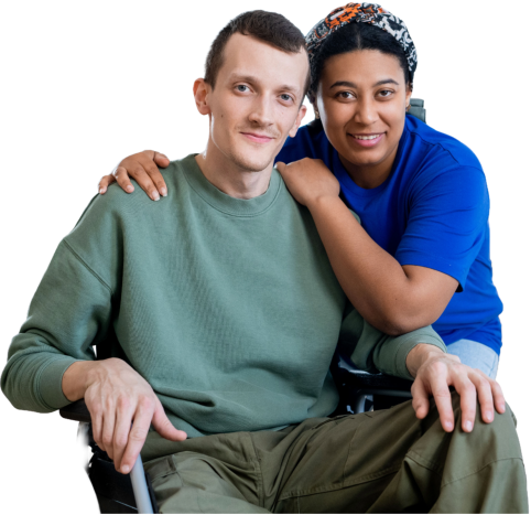 A woman with patterned head covering is standing behind a man in wheelchair, looking at the camera. Her arm is around him. In the background two people are working on assembling a chair in the bright, airy living room. This image is perfect for health and wellness projects, social connection, and home improvement. The friendly, caring mood will make the viewer feel supported and connected.