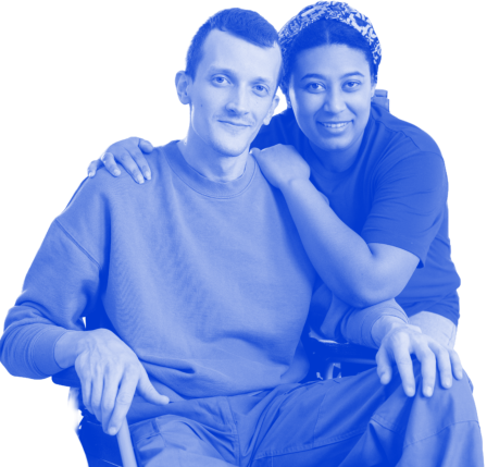 A woman with patterned head covering is standing behind a man in wheelchair, looking at the camera. Her arm is around him. In the background two people are working on assembling a chair in the bright, airy living room. This image is perfect for health and wellness projects, social connection, and home improvement. The friendly, caring mood will make the viewer feel supported and connected.