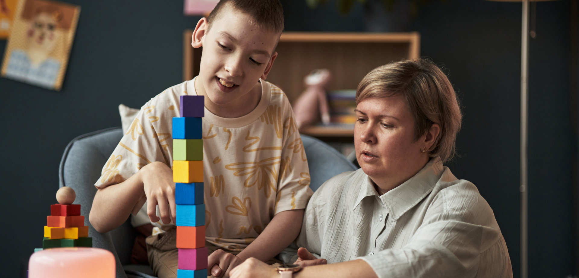 A child and an adult are playing with colorful wooden blocks at a wooden table in a home setting. The child is constructing a tall tower with the blocks, while the adult watches with their hands on the table. Other wooden blocks are placed around the table. Natural lighting and a playful mood make this image perfect for educational and family themed projects.