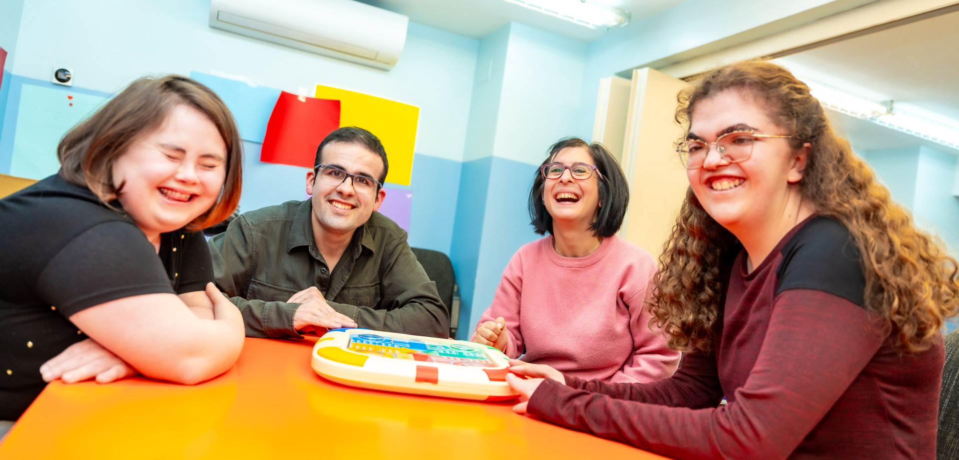 Four cheerful adults are gathered around a bright orange table indoors, engaging in a playful learning activity. They are all smiling and appear to be enjoying themselves. The setting looks like a classroom or community center, with light blue walls and an air conditioning unit. The photo's bright lighting and happy mood makes it suitable for projects about education, adult learning, collaboration, or community engagement.