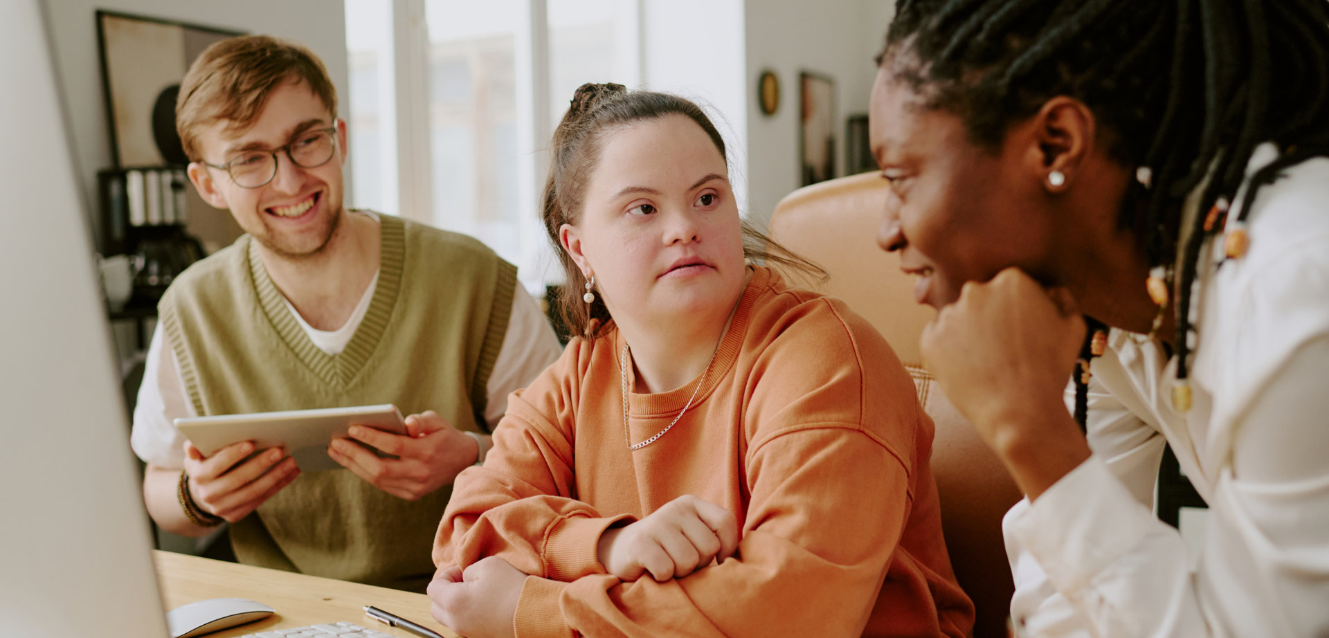 In a brightly lit office setting, three individuals are gathered around a wooden desk, engaged in a collaborative meeting. A man with glasses and a light-colored sweater vest is holding a tablet and smiling. A woman in an orange sweater looks toward another woman in a white blouse, who has her chin resting on her hand. The desk is equipped with a computer monitor, keyboard, mouse, open notebook and other office supplies. This image evokes collaboration, technology, and workplace dynamics and would be suitable for projects related to workplace inclusion, technology, or corporate environments.