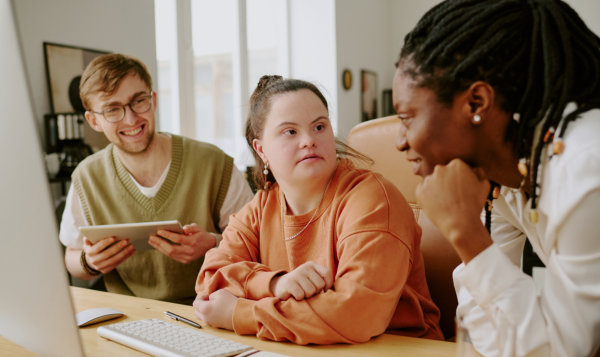 In a brightly lit office setting, three individuals are gathered around a wooden desk, engaged in a collaborative meeting. A man with glasses and a light-colored sweater vest is holding a tablet and smiling. A woman in an orange sweater looks toward another woman in a white blouse, who has her chin resting on her hand. The desk is equipped with a computer monitor, keyboard, mouse, open notebook and other office supplies. This image evokes collaboration, technology, and workplace dynamics and would be suitable for projects related to workplace inclusion, technology, or corporate environments.