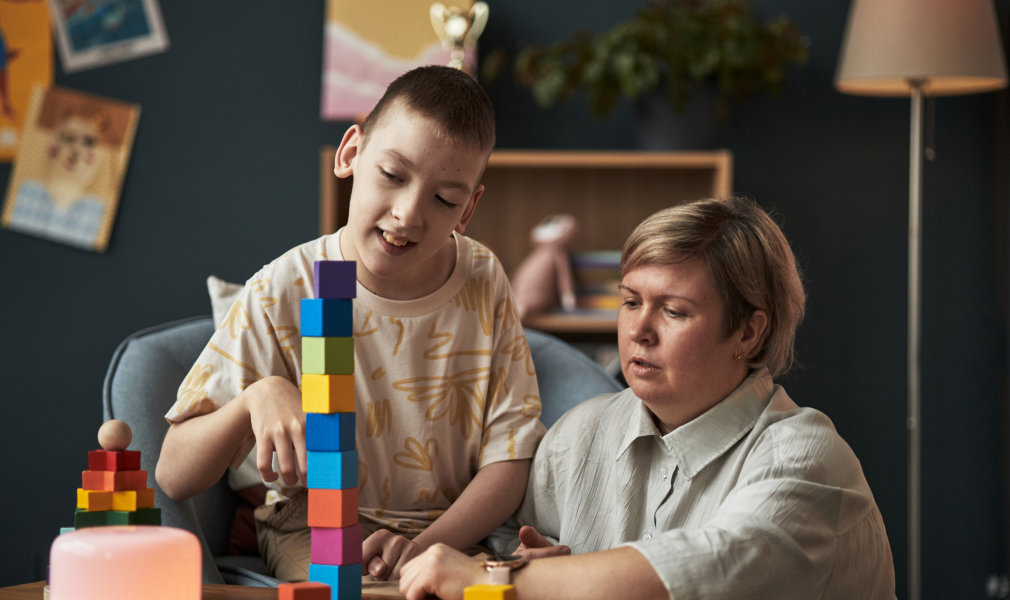 A child and an adult are playing with colorful wooden blocks at a wooden table in a home setting. The child is constructing a tall tower with the blocks, while the adult watches with their hands on the table. Other wooden blocks are placed around the table. Natural lighting and a playful mood make this image perfect for educational and family themed projects.