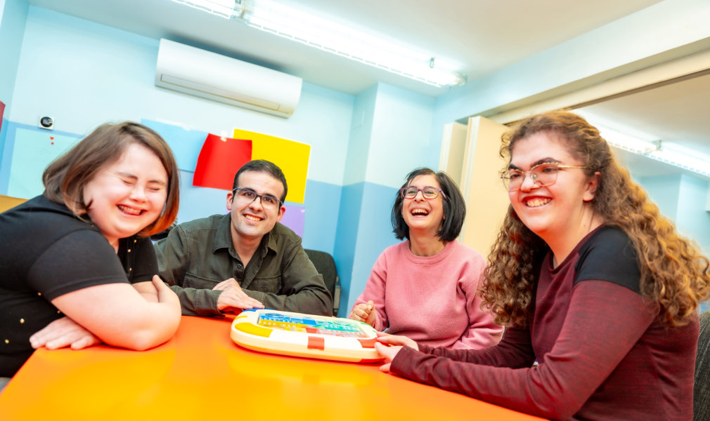 Four cheerful adults are gathered around a bright orange table indoors, engaging in a playful learning activity. They are all smiling and appear to be enjoying themselves. The setting looks like a classroom or community center, with light blue walls and an air conditioning unit. The photo's bright lighting and happy mood makes it suitable for projects about education, adult learning, collaboration, or community engagement.