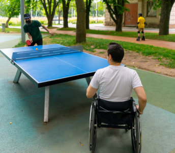 Two men playing table tennis on an outdoor court with another person visible in the background. One man with short dark hair and a white t-shirt is in a wheelchair. The other man with dark hair and a beard, wearing a dark shirt, is getting ready to hit the orange ball. The court is blue and black. There is an individual on roller blades in the background. The photo evokes a sense of an active lifestyle and accessibility, perfect for projects centered on sports, recreation, and inclusive activities.